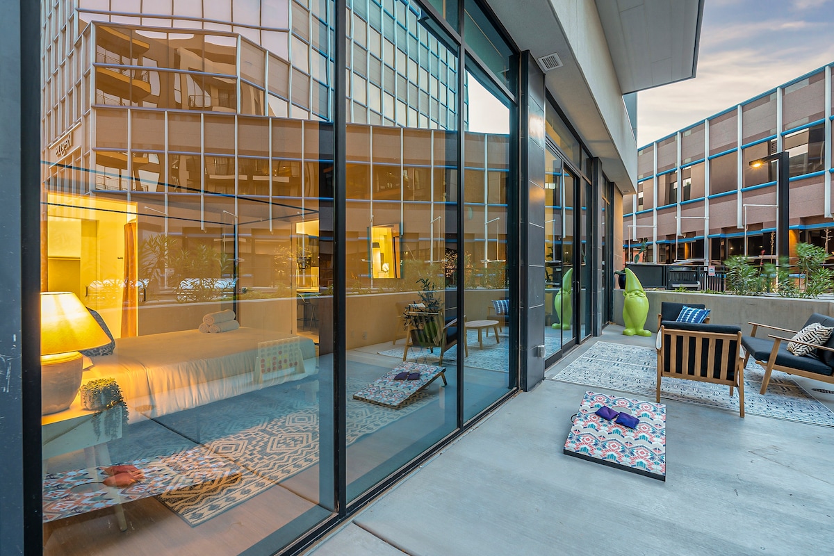 An outdoor balcony area features large glass doors that provide a view of the interior space. Colorful floor cushions are arranged on a patterned rug, alongside wooden seating and green decorative accents, creating a relaxed atmosphere with reflections of the surrounding buildings.