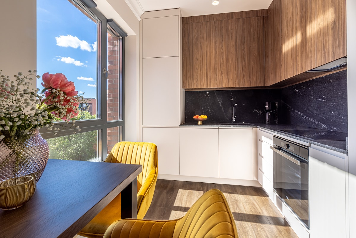 A modern kitchen is featured with sleek cabinetry and a dark stone countertop. Bright, natural light fills the space through large windows. A small dining table is accompanied by two yellow upholstered chairs. A vase of flowers adds a touch of color to the setting.