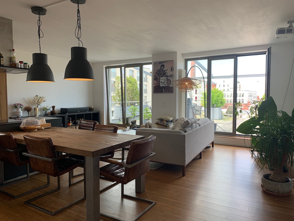 An open living area features a large wooden dining table surrounded by four leather chairs. A comfortable sofa is positioned beside expansive windows that allow natural light to fill the space, while a decorative plant adds a touch of greenery.