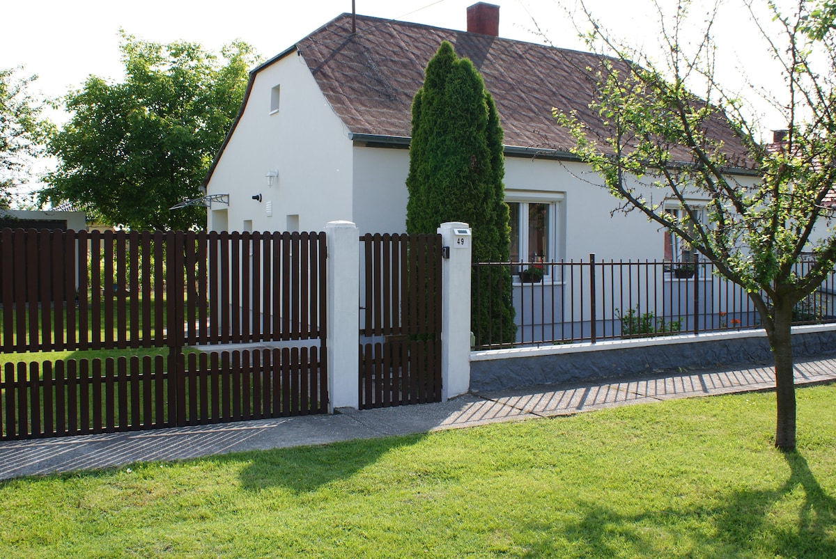 A welcoming house is presented with a simple exterior, featuring a well-maintained garden. A wooden fence and gate mark the entrance, providing a sense of privacy. Greenery surrounds the house, enhancing the tranquil environment.