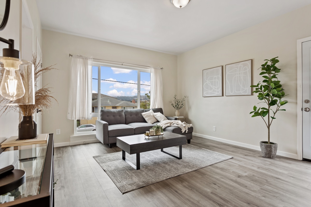 A bright living area features a gray sofa positioned near a large window that allows natural light to enter. A coffee table sits on a textured area rug, complemented by decorative plants and minimalist wall art. Light curtains frame the window, enhancing a spacious feel.