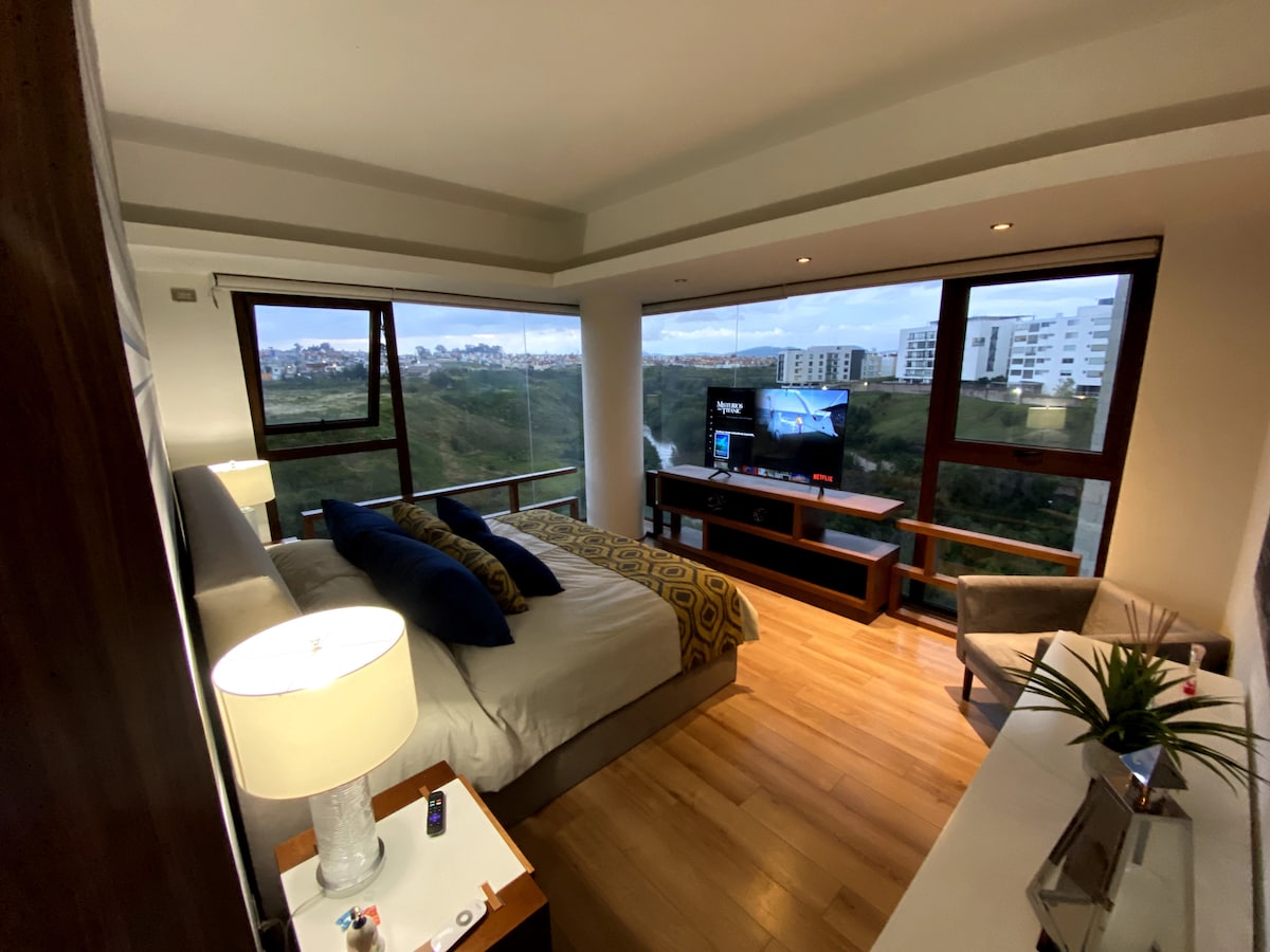 A well-appointed bedroom features a spacious bed adorned with decorative pillows, accompanied by a stylish lamp beside it. A television is mounted on a moderate entertainment center, while large windows provide ample natural light and a view of the surrounding landscape.