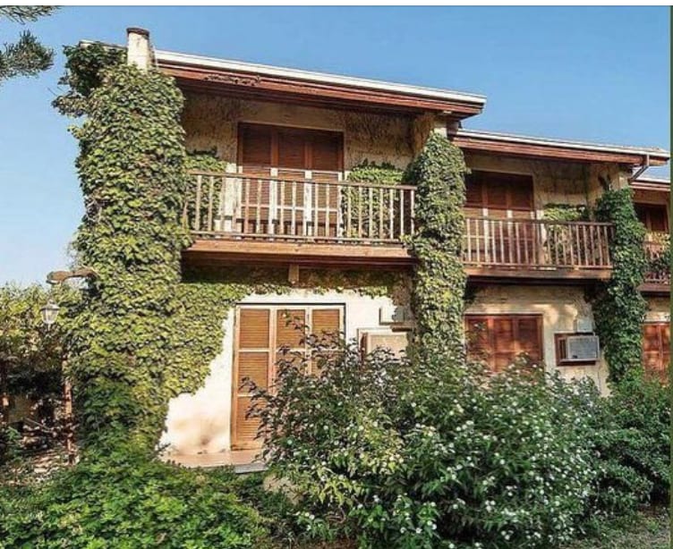 A two-story bungalow is enveloped in lush green vines, with wooden balconies featuring slatted railings. Large windows allow natural light to illuminate the space. Flowering bushes frame the entrance, while the clear blue sky provides a serene backdrop to the inviting structure.