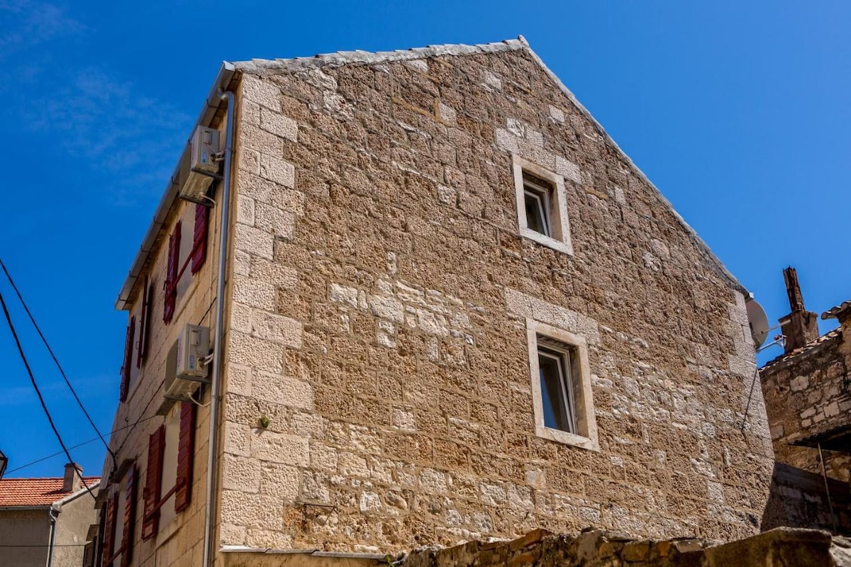 The exterior of a recently renovated Dalmatian stone house is displayed, showcasing the natural texture of the stone. The building features a sloped roof and several windows, framed by red shutters. A clear blue sky serves as the backdrop.