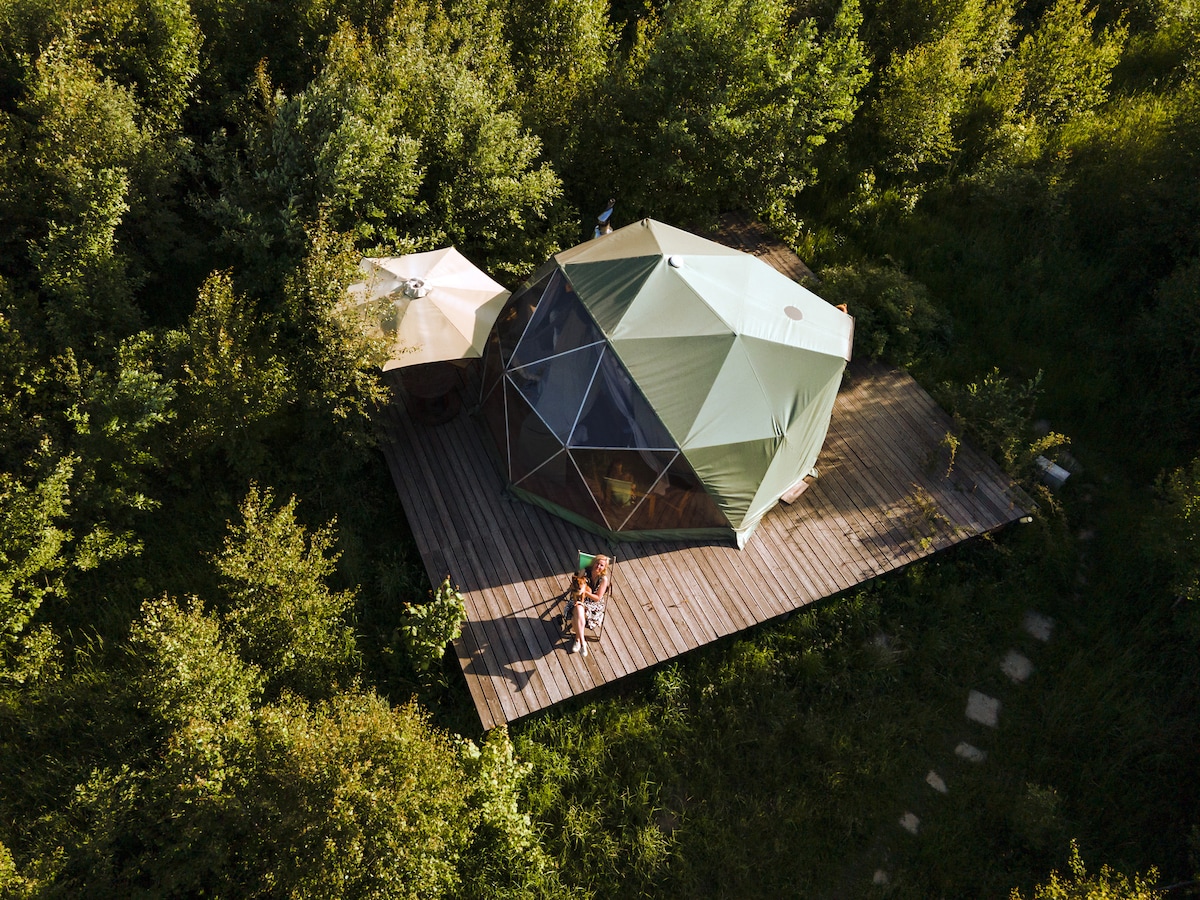 A spherical tent is nestled on a wooden deck surrounded by lush greenery. A shaded sitting area with a table and chair offers space for relaxation, while pathways lead through the vibrant meadow. The tent's geometric design complements the natural setting.