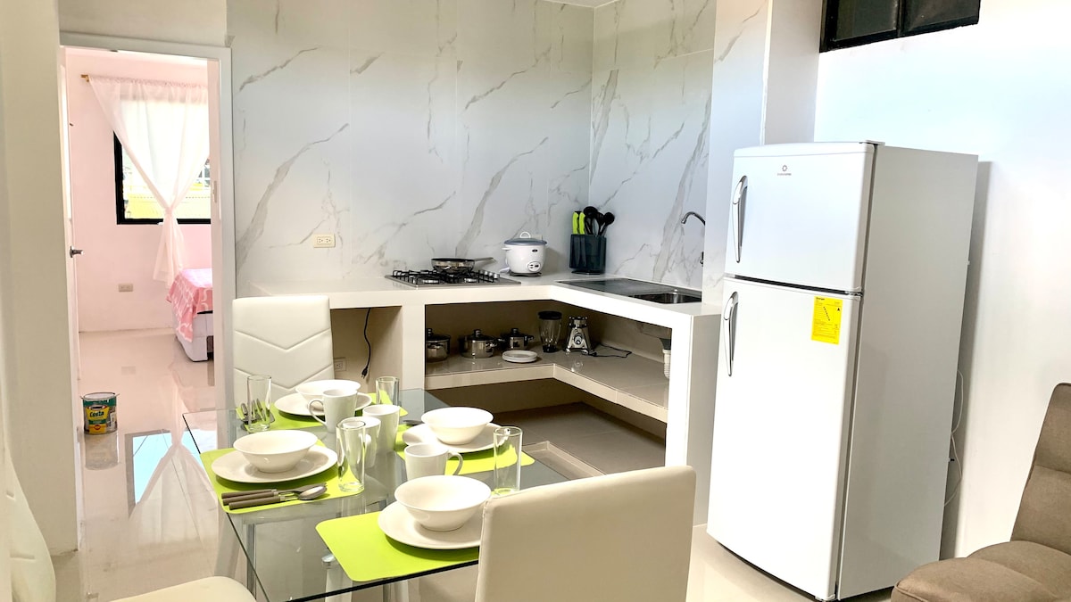 A bright kitchen and dining area showcases sleek white surfaces and modern appliances. A glass dining table is set for four, featuring dinnerware and silverware. The kitchen area includes a gas stove, refrigerator, and essential cooking utensils, creating a functional and inviting space.