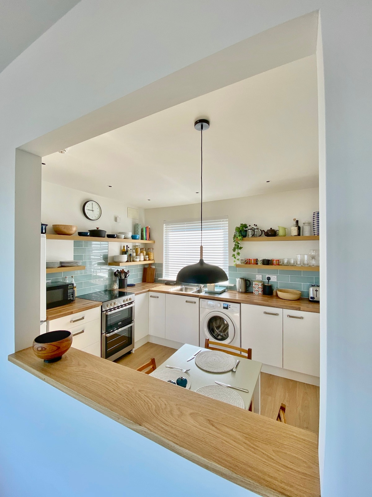 The kitchen features modern cabinetry and open shelving stocked with a variety of kitchenware. A circular dining table with wooden chairs is positioned under a pendant light, providing an inviting space for meals. Large windows offer natural light, enhancing the bright atmosphere.