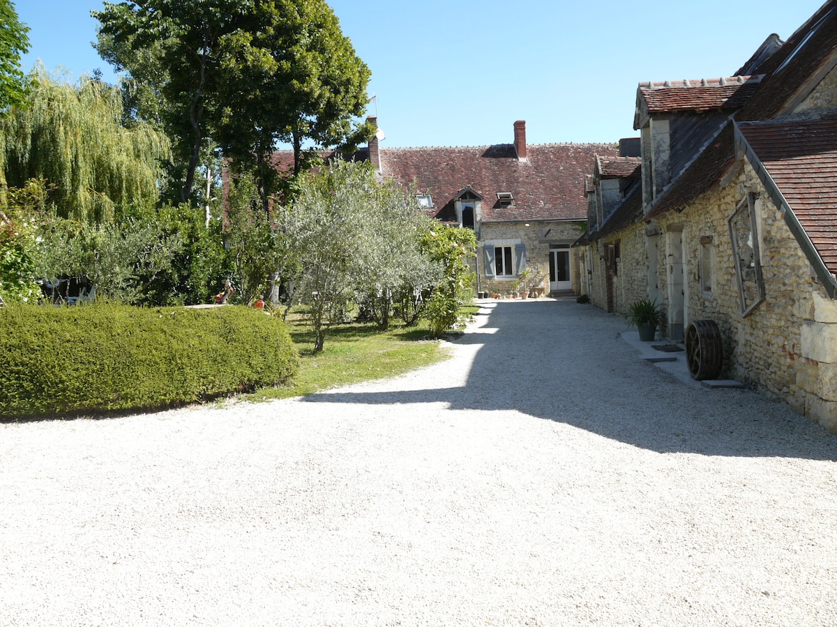 A spacious gravel courtyard is surrounded by lush greenery and trees, with a charming stone building to the right. The sun casts soft shadows, creating a serene outdoor setting inviting relaxation.