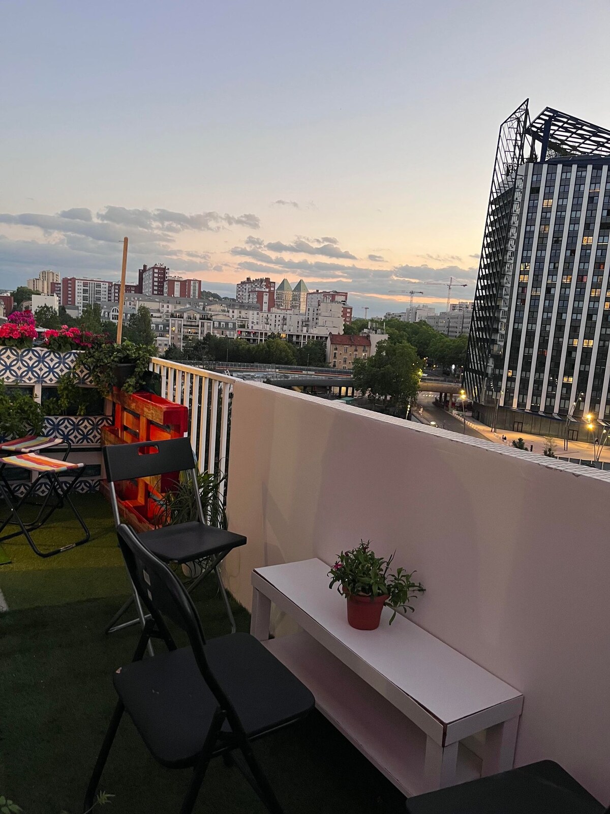 A balcony area is displayed with several black chairs and a small table. Potted plants are placed on the table, while colorful planters are visible along the railing. The surrounding cityscape and buildings are seen against a sunset sky, creating a relaxed outdoor space.