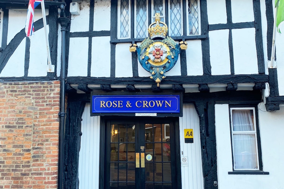 The entrance of the Rose and Crown hotel is framed by traditional black and white timber framing. A royal crest is prominently displayed above the door, with the hotel name 'Rose & Crown' featured in blue lettering. Flags are visible on either side, adding to the establishment's charm.