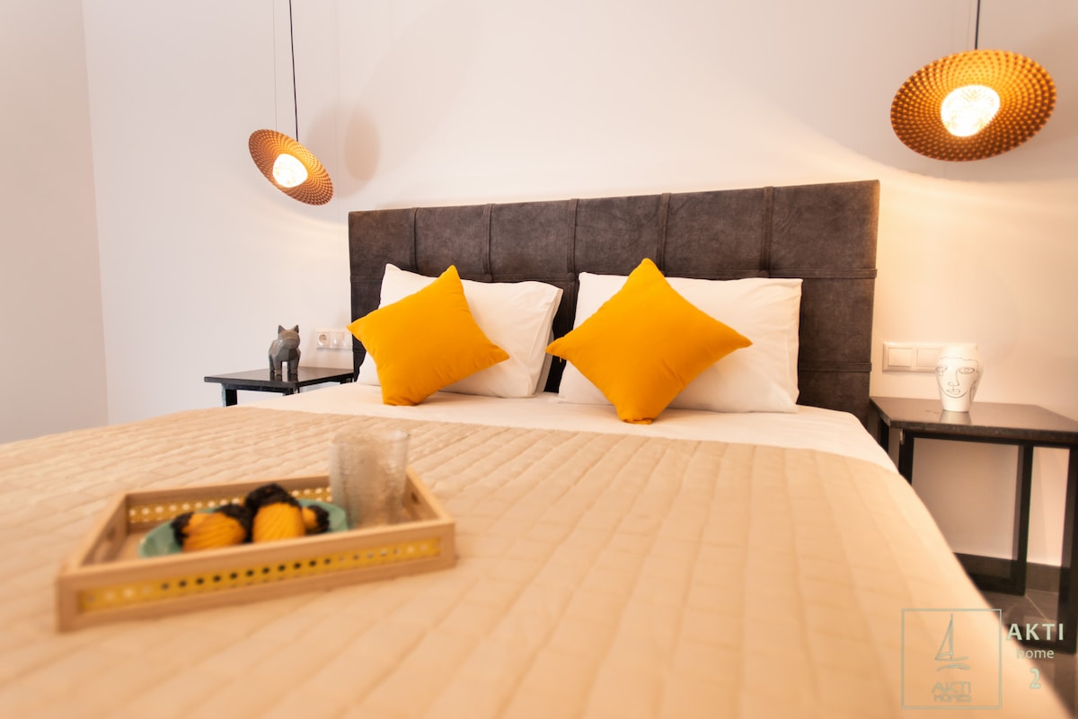 A modern bedroom showcases a spacious bed dressed in neutral tones, accented by two yellow pillows. Stylish pendant lights hang above, illuminating the space. A wooden tray with decorative items is placed on the bed, while a small sculpture adds character to the room.