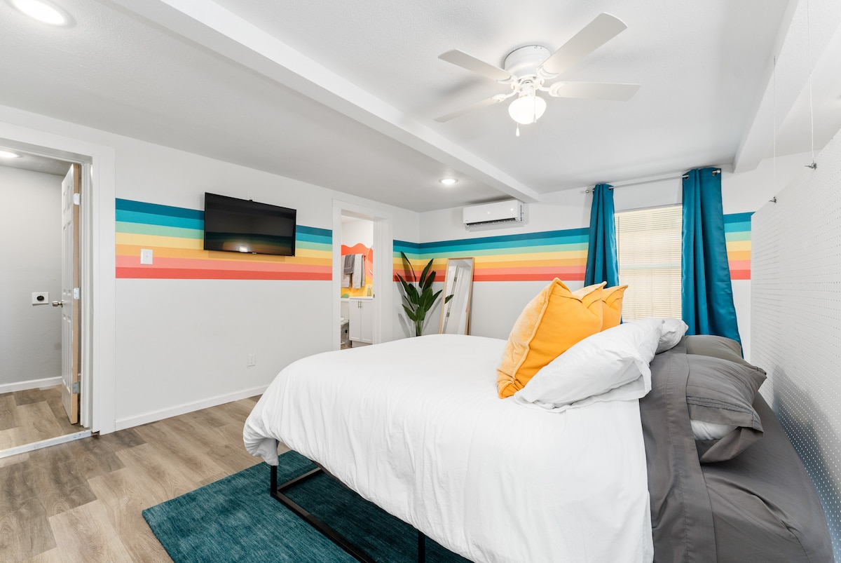 A comfortable sleeping space features a spacious bed with layered bedding. The colorful accent wall displays horizontal stripes in vibrant hues. Natural light filters through the window, illuminating the room. A potted plant adds a touch of greenery, while a ceiling fan enhances air circulation.