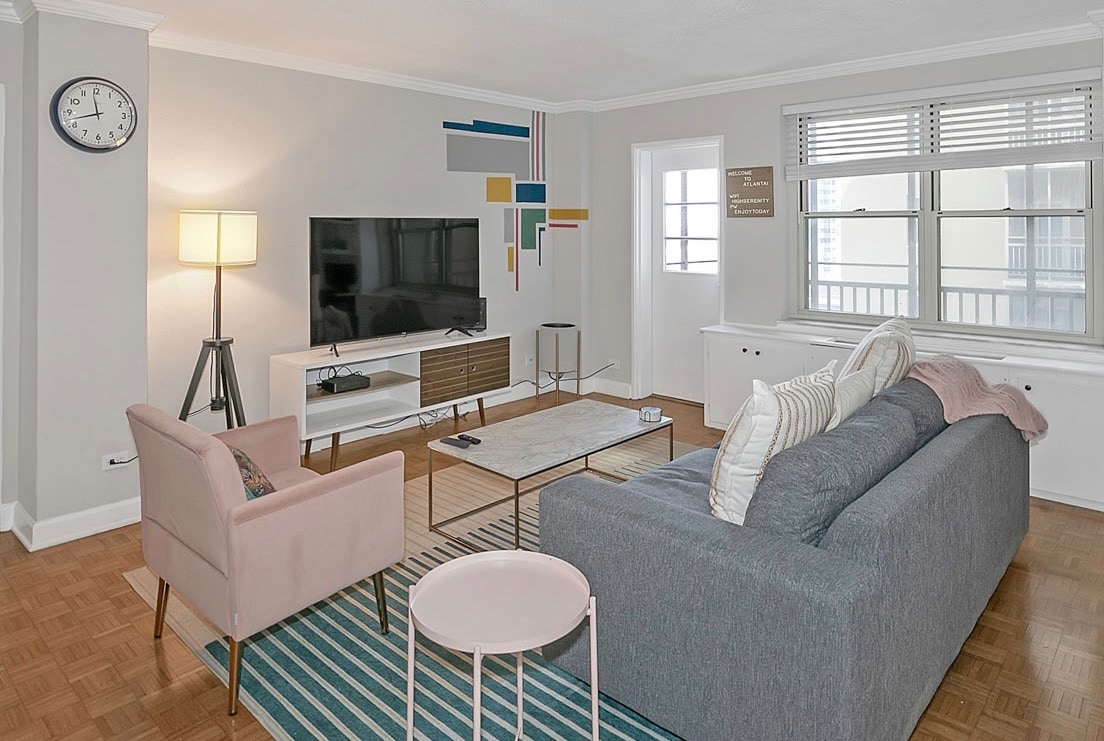 A living room area features a modern design with a large sofa and an armchair. A light-colored coffee table is positioned on a patterned rug. A flat-screen TV is mounted on the wall opposite the sofa, while a clock and decorative accents add interest to the space.
