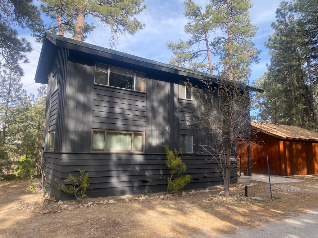 The exterior of the home displays a modern black facade, complemented by large windows that invite natural light. Surrounding pines enhance the serene setting, while a wooden structure sits nearby, presenting a rustic contrast. A gravel path leads to the entrance, emphasizing the tranquil environment.