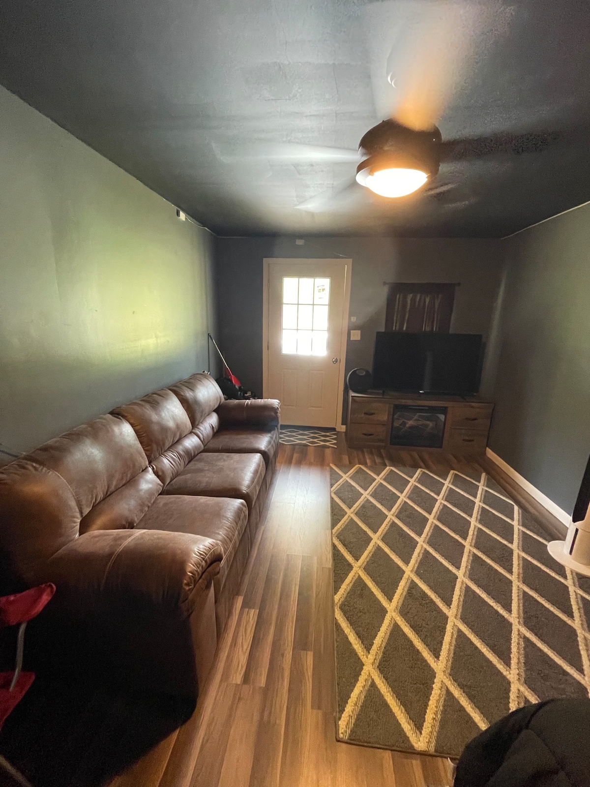 A cozy living area features a brown leather sofa positioned against a dark wall. Light filters through a door with multiple panes of glass, leading to the exterior. A striped rug enhances the warm tones of the space, while a TV is mounted on a wooden stand.