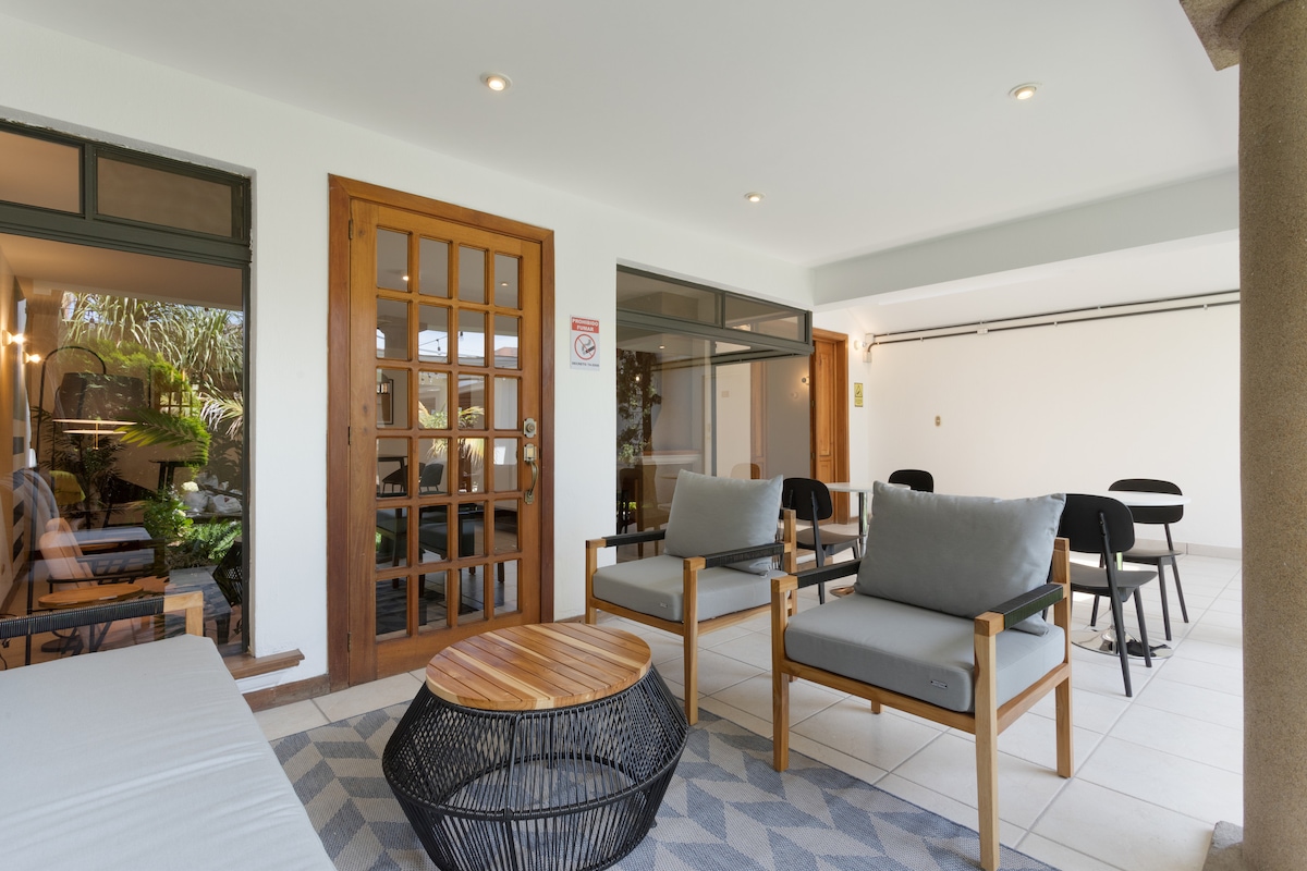A communal lounge area features comfortable seating with light grey cushions on wooden frames, complemented by a round wooden table. Large windows provide natural light, while a modern touch is added by the patterned rug and the greenery visible through the glass.