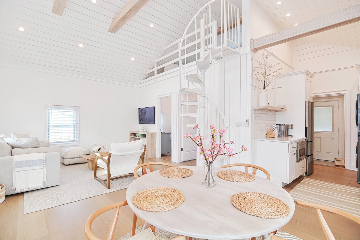 An airy living area is highlighted by a spiral staircase leading to a loft space. Light-colored walls and furnishings complement warm wood accents. A round dining table is set with woven placemats, while a cozy seating area features a television and large windows allowing natural light to fill the room.