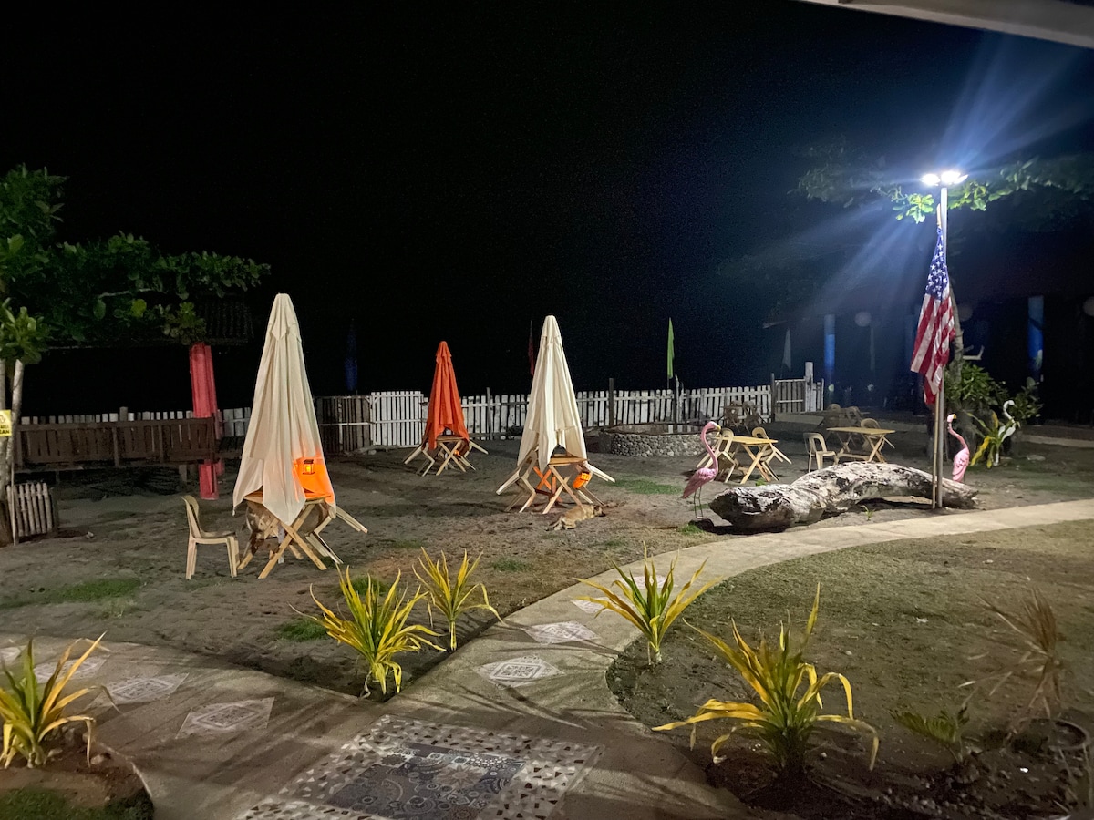 A serene outdoor area is illuminated at night, featuring several wooden chairs arranged under large umbrellas. The pathway is bordered by ornamental plants, with a flag visible nearby. A peaceful atmosphere is created by the soft lighting around the space.