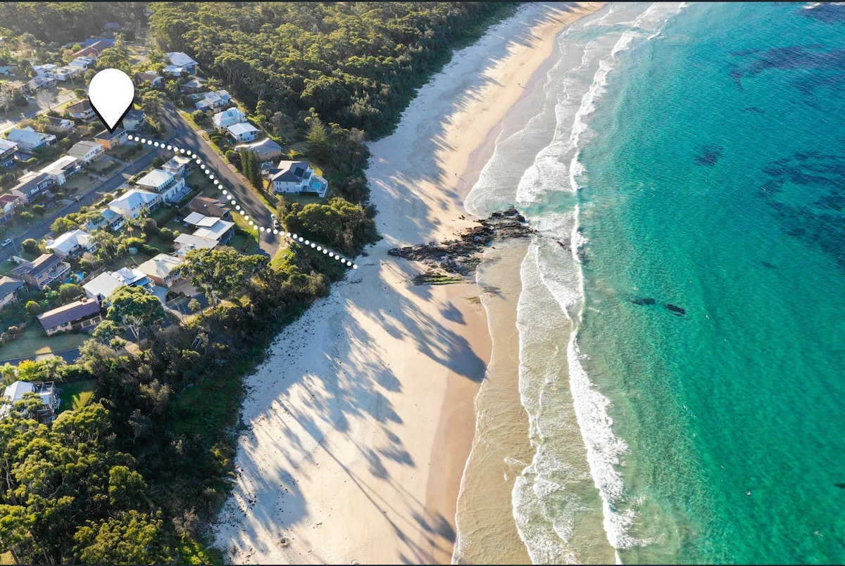 An aerial view captures the scenic coastline, displaying gentle waves lapping against the sandy beach. The shoreline is flanked by lush greenery and residential structures, with a clear path leading from the houses to the water's edge.