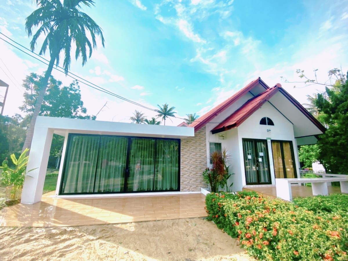 A single-story bungalow is shown, featuring a sloped red roof and large glass windows. A spacious outdoor area is visible, with a tiled patio and green foliage. Bright natural light streams through the windows, enhancing the welcoming appearance of the home.