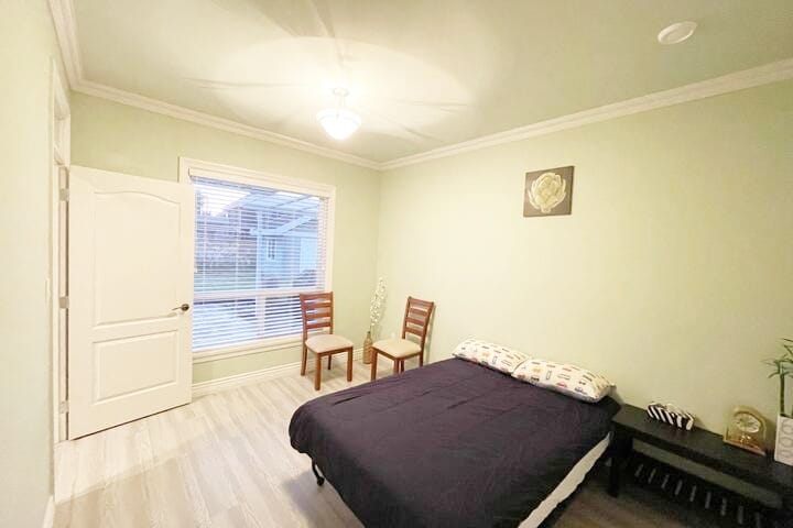 The square bedroom features a spacious bed with a dark blanket and colorful pillows. Two wooden chairs are positioned near the window, which allows natural light to fill the room. A simple wall decoration adds a touch of design to the green walls.