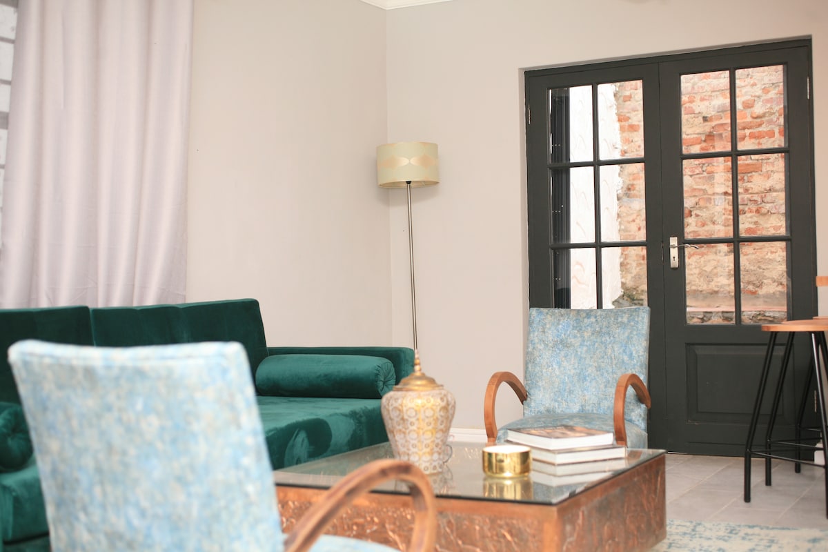 A comfortable living area features a green sofa and two blue accent chairs. A glass coffee table sits in the center, adorned with decorative items and books. Natural light enters through a door with multiple panes, revealing a textured brick wall in the background.