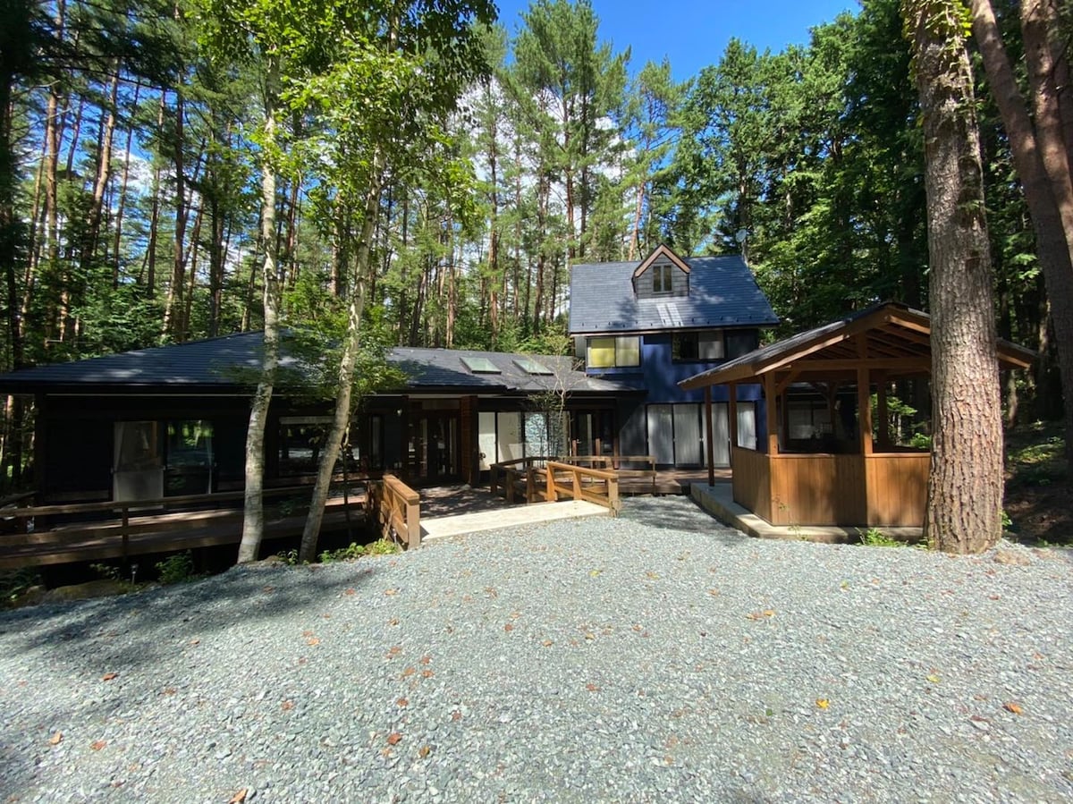 A spacious cottage is nestled among tall trees, featuring a dark roof and light wood accents. The outdoor area includes a wooden deck leading to a covered seating space, set against a backdrop of natural greenery and a pebbled driveway.