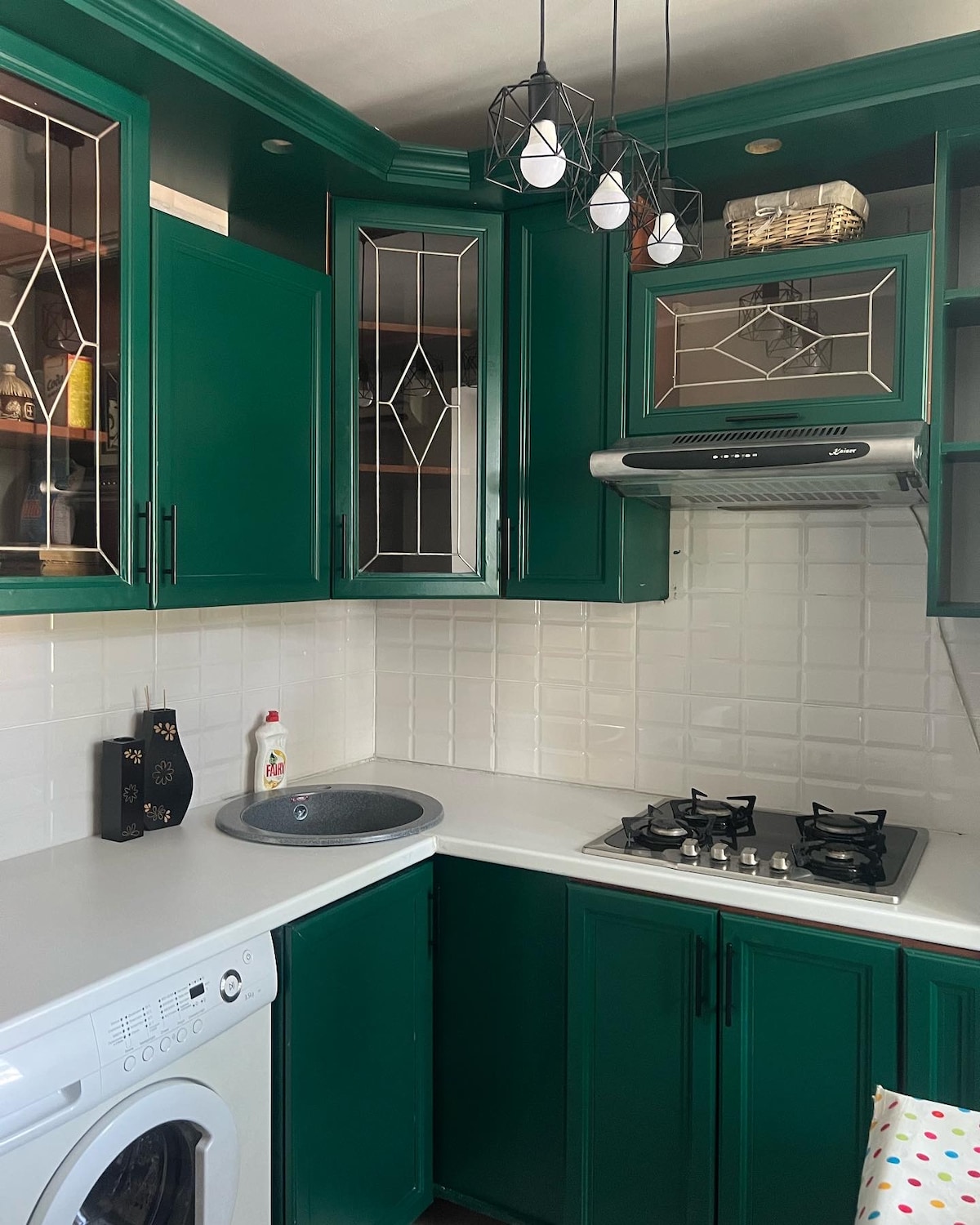 The kitchen features striking green cabinetry with glass accents, providing a modern and stylish look. A gas stove with a vent hood is situated beside a small sink, while a washing machine is integrated into the design. Bright lighting is offered by hanging fixtures.