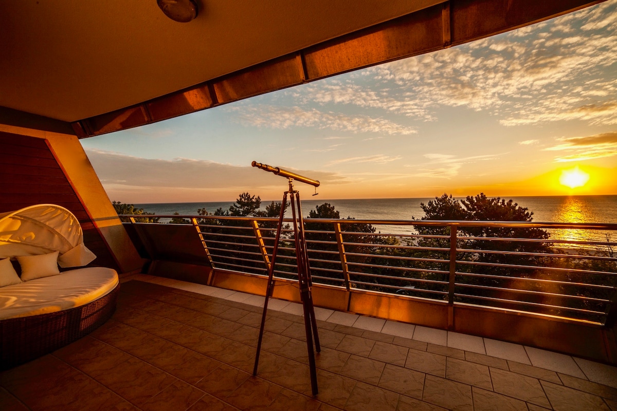 A spacious terrace offers a direct view of the sea at sunset. A telescope is positioned to enhance the scenic vista. Soft lounge seating is present, and the warm tones of the sunset create a serene atmosphere against the expansive ocean backdrop.