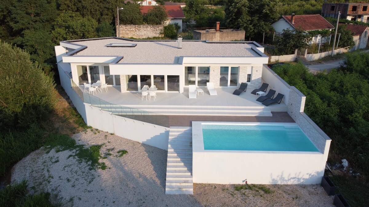 An aerial view reveals a modern villa with a sleek white exterior and large glass doors, surrounded by lush greenery. An inviting outdoor pool is situated beside a spacious terrace, furnished with lounge chairs and dining tables. The landscape showcases a blend of natural and built environments.