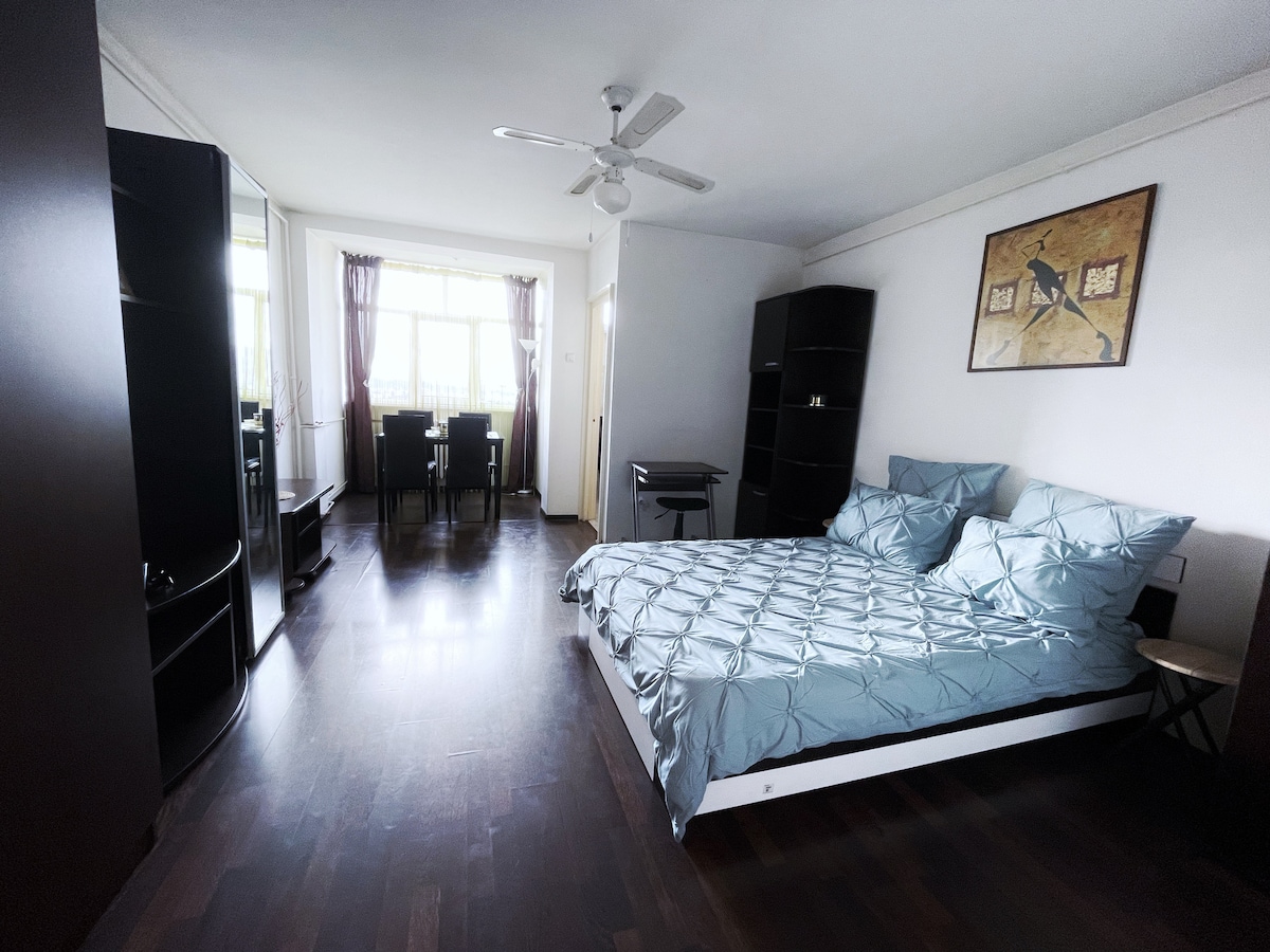 A spacious studio is showcased, featuring a large bed with a light blue quilt and matching pillows. Adjacent, a dining area with a table and four chairs is visible. Natural light filters in through floor-to-ceiling windows, highlighting the dark wooden flooring.