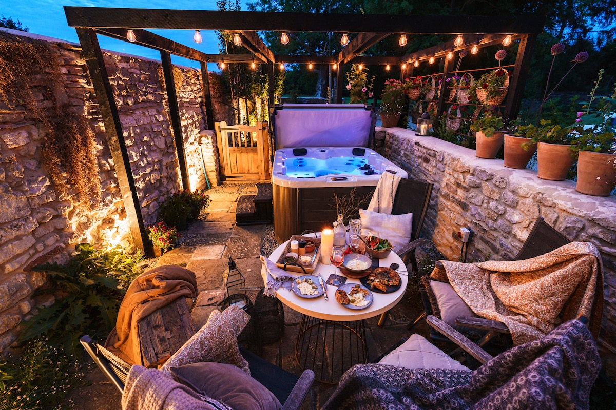 An al-fresco patio area features a private hot tub surrounded by potted plants and soft lighting. A table is set with various dishes, and cozy seating options are arranged nearby, inviting relaxation and comfort in a picturesque outdoor setting.