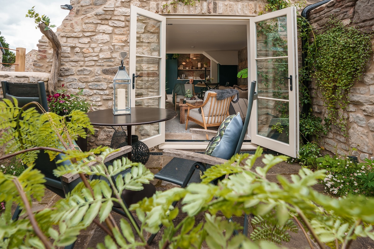 Outdoor seating is provided on a patio area, featuring a round table and relaxed chairs. Open French doors reveal a cozy interior with plush seating and greenery. The stone wall offers a rustic charm, while potted plants frame the scene, enhancing the peaceful atmosphere.