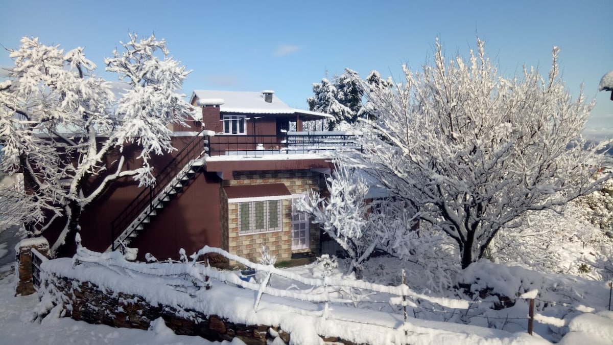 A homestay nestled in nature is surrounded by snow-covered trees and a blanket of white snow. The exterior features a wooden balcony and a staircase leading to an upper level, with large windows offering views of the winter landscape.