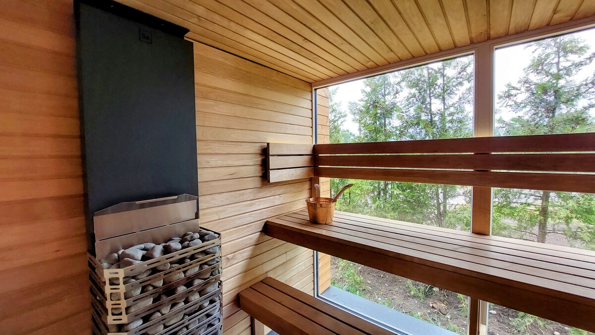 A wooden sauna interior features two spacious benches along opposing walls, constructed from smooth timber. A metal stove is situated in one corner, surrounded by a stack of stones. Large windows provide a view of the greenery outside, enhancing the serene atmosphere.