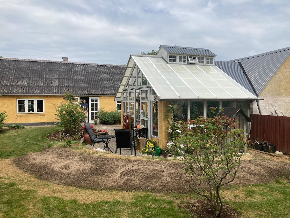 A charming orangery is surrounded by a well-maintained garden, featuring a mix of greenery and flowering plants. Outdoor seating arrangements are visible, inviting relaxation. The structure showcases large glass panels, allowing natural light to fill the interior, while a rustic yellow building is seen in the background.