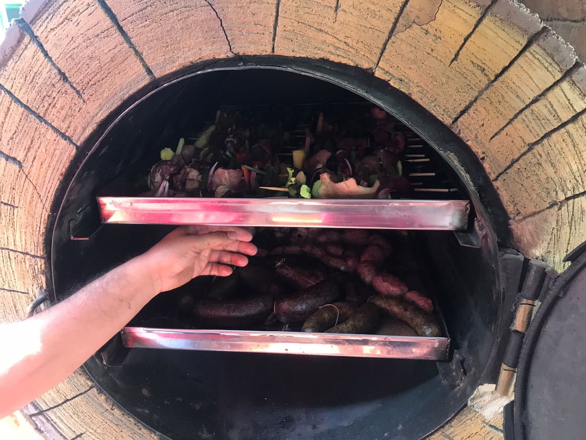 An outdoor oven is showcased, featuring two trays filled with various sausages and vegetables. The brick archway surrounds the oven's entrance, with a hand visible, inserting ingredients for cooking. The interior is designed for grilling, emphasizing a communal cooking experience.