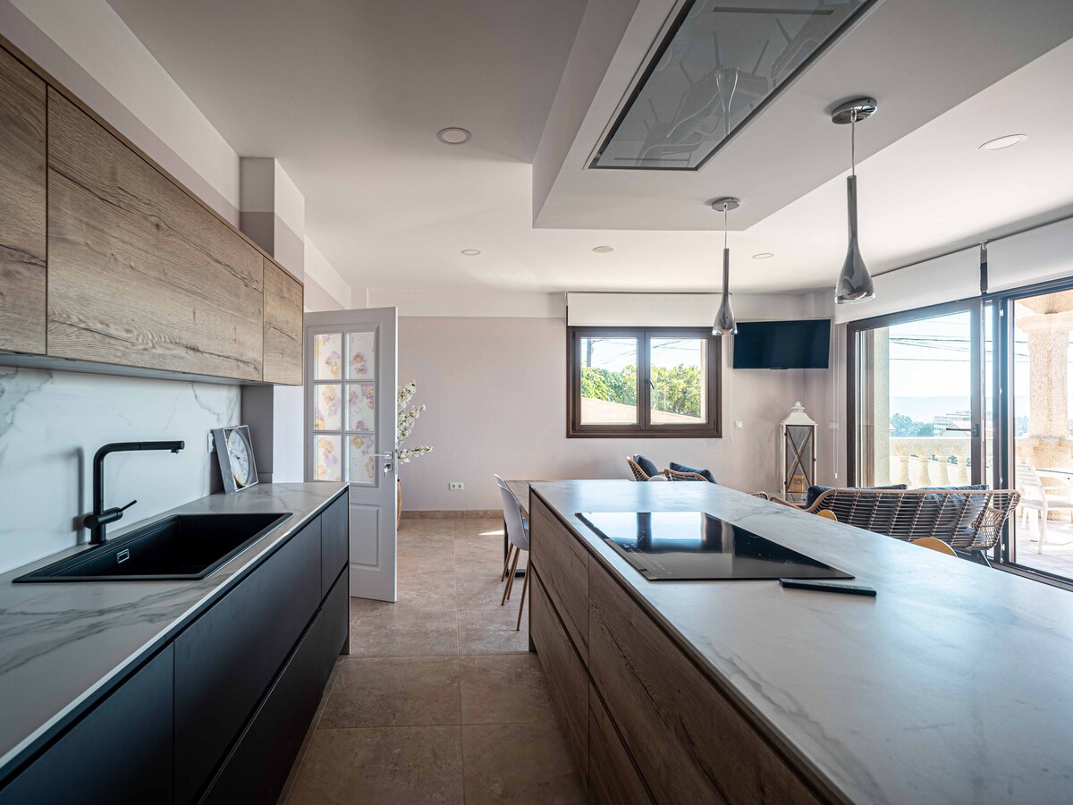 A modern kitchen features sleek wooden cabinetry and a large island with a cooktop. Natural light fills the space through a large window, illuminating the open layout that connects to a dining area. Contemporary pendant lights hang above, enhancing the minimalist design.