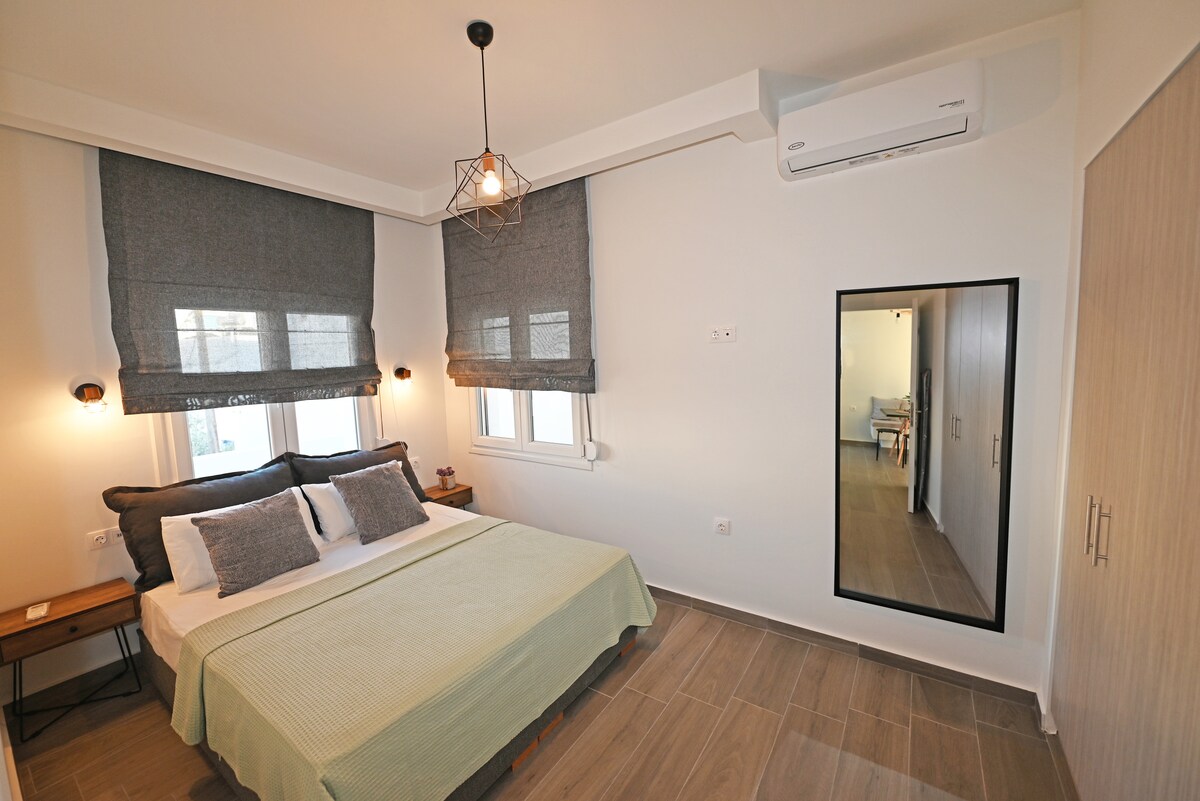 A modern bedroom features a spacious bed with neutral bedding, flanked by bedside tables and lamps. Large windows are dressed with gray shades, allowing natural light to filter in. A full-length mirror is mounted on the wall, contributing to the room's bright and airy feel.