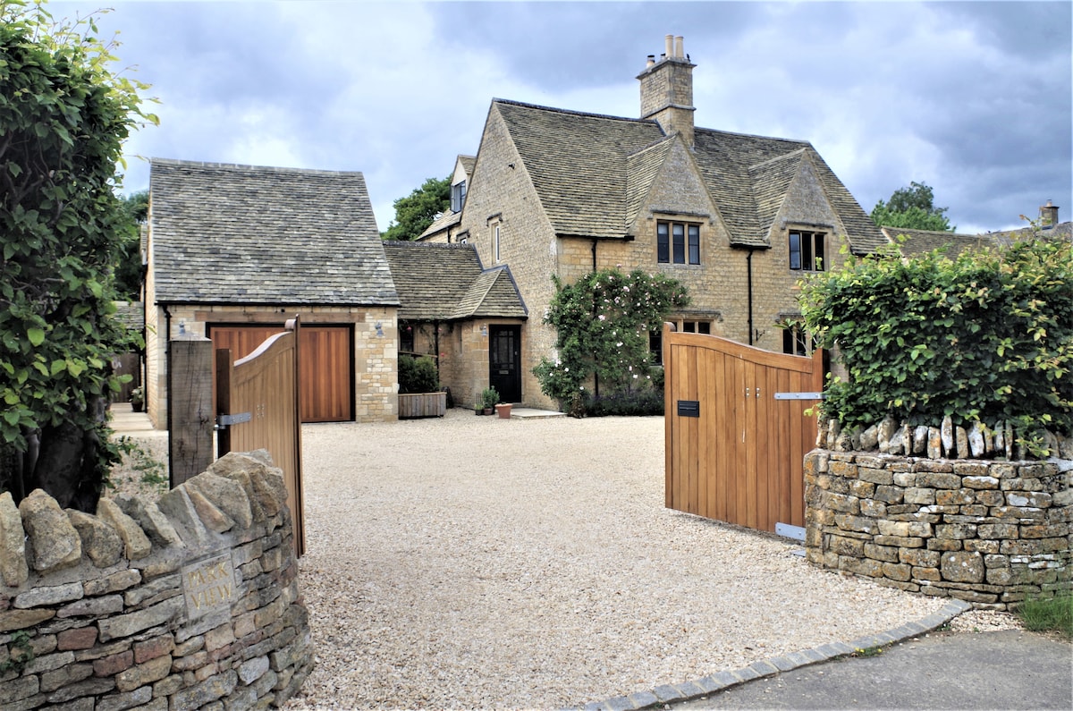 A stone cottage with traditional Cotswold architecture is displayed beneath a cloudy sky. The property is framed by wooden gates opening onto a spacious gravel courtyard, with lush greenery surrounding the entrance and stone wall.