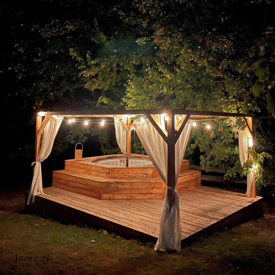 An outdoor jacuzzi area is accentuated by warm lighting, creating a calming ambiance. Wooden decking surrounds the jacuzzi, while sheer curtains drape from the pergola structure. Natural foliage frames the space, offering a sense of privacy and seclusion.