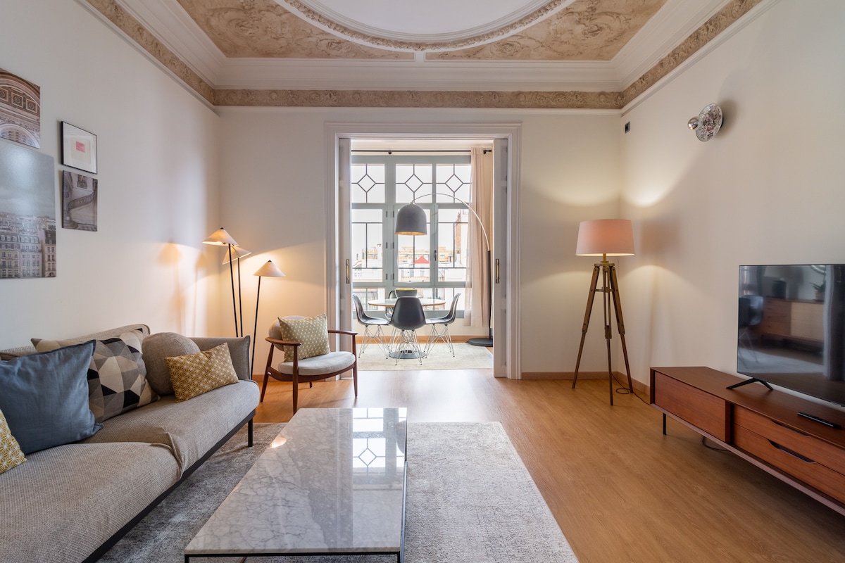 A spacious living room features a comfortable, curvy sofa with decorative pillows, accompanied by stylish floor lamps. A glass coffee table sits on a soft area rug, while natural light filters in from the open doorway, highlighting a modern dining area beyond.
