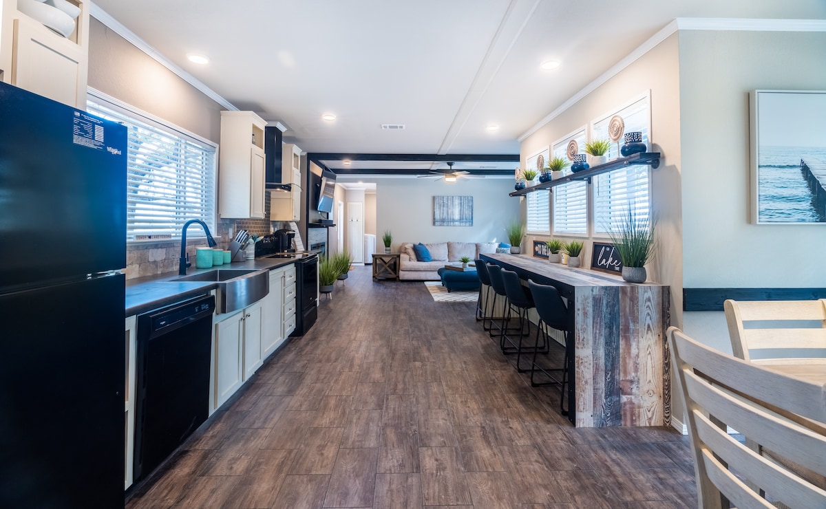 An open living and kitchen area features modern appliances and a spacious countertop bar with casual seating. Sunlight streams through multiple windows, illuminating the space. The living area includes a comfortable couch and decorative plants, creating an inviting space for gathering and relaxing.