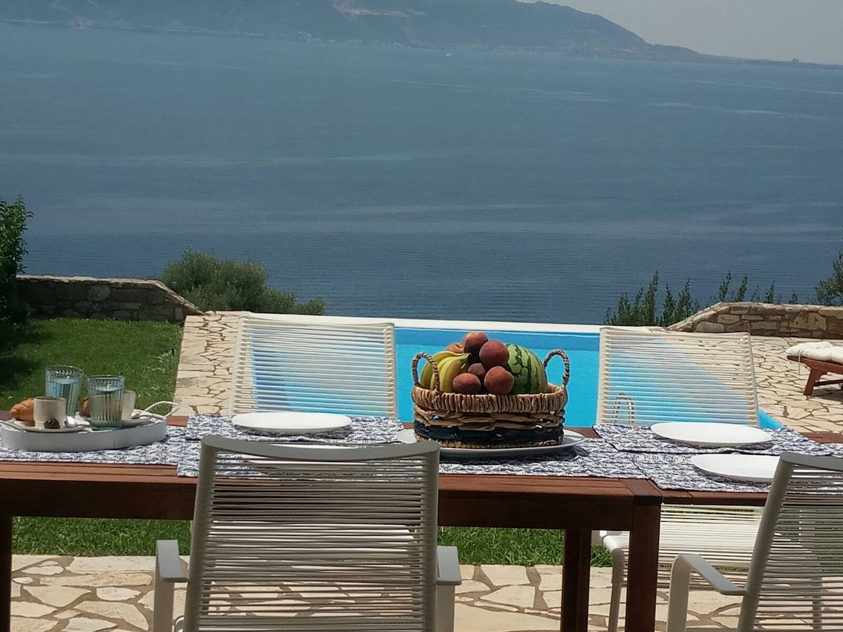 An outdoor dining area is set with a table adorned with a colorful fruit basket, placing an emphasis on a picturesque view of the sea. Sun loungers are positioned near a sparkling pool, while a stone patio connects with the surrounding greenery.