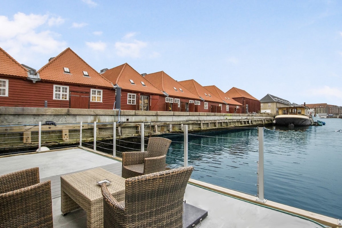 A seating area is presented on a spacious deck overlooking a calm canal. Two woven chairs and a small table are positioned near the water, while colorful buildings with red roofs line the opposite shore, reflecting in the clear water.