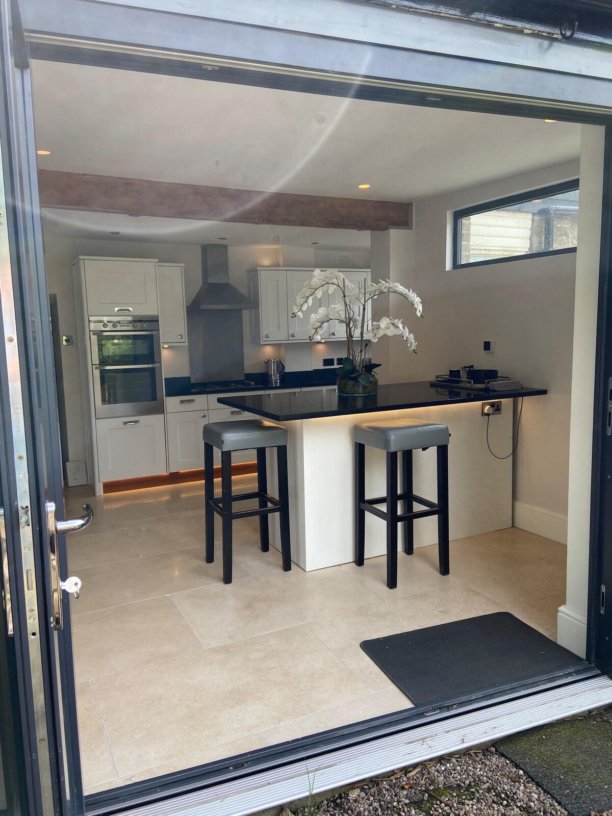 The open-plan kitchen features a central island with two high stools, complemented by contemporary white cabinetry and modern appliances. Natural light enters through a large door and window, enhancing the bright and functional cooking space.