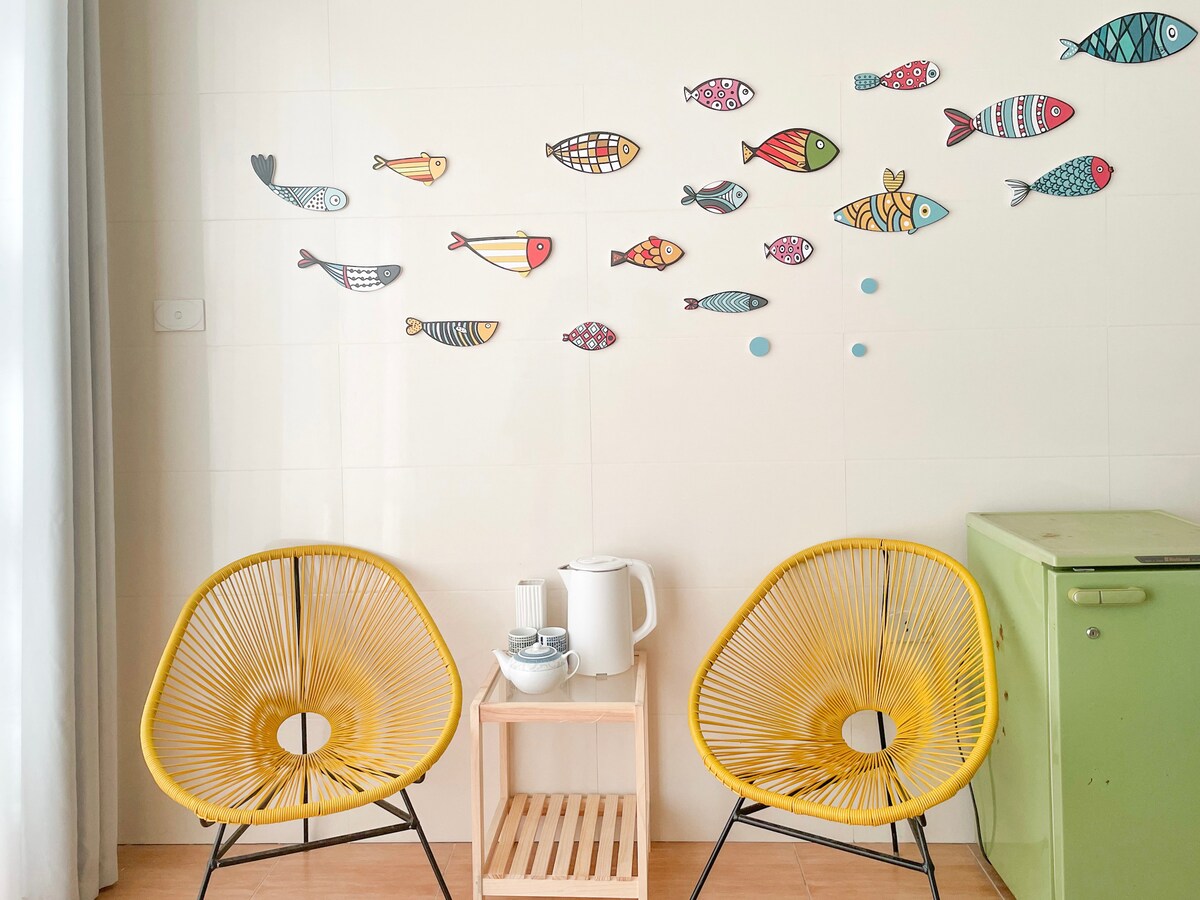 Two vibrant yellow chairs are positioned beside a wooden side table, which holds a teapot and cups. The wall is adorned with colorful fish illustrations of various sizes, creating a playful and cheerful atmosphere. A small green refrigerator is visible in the corner.