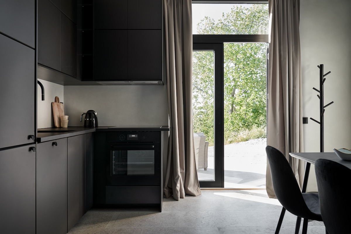 A modern kitchen area features sleek black cabinetry, an oven, and a kettle. Large glass doors open to an outdoor space with trees nearby. Soft curtains frame the windows, allowing natural light to enhance the minimalist design.