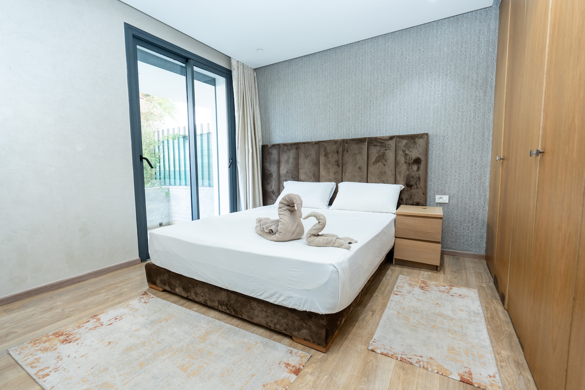 A spacious bedroom is featured, with a comfortable bed and a modern headboard. Natural light enters through large glass doors that lead to an outdoor area. A soft rug lies beneath the bed, complementing the warm wood tones of the flooring and furniture.