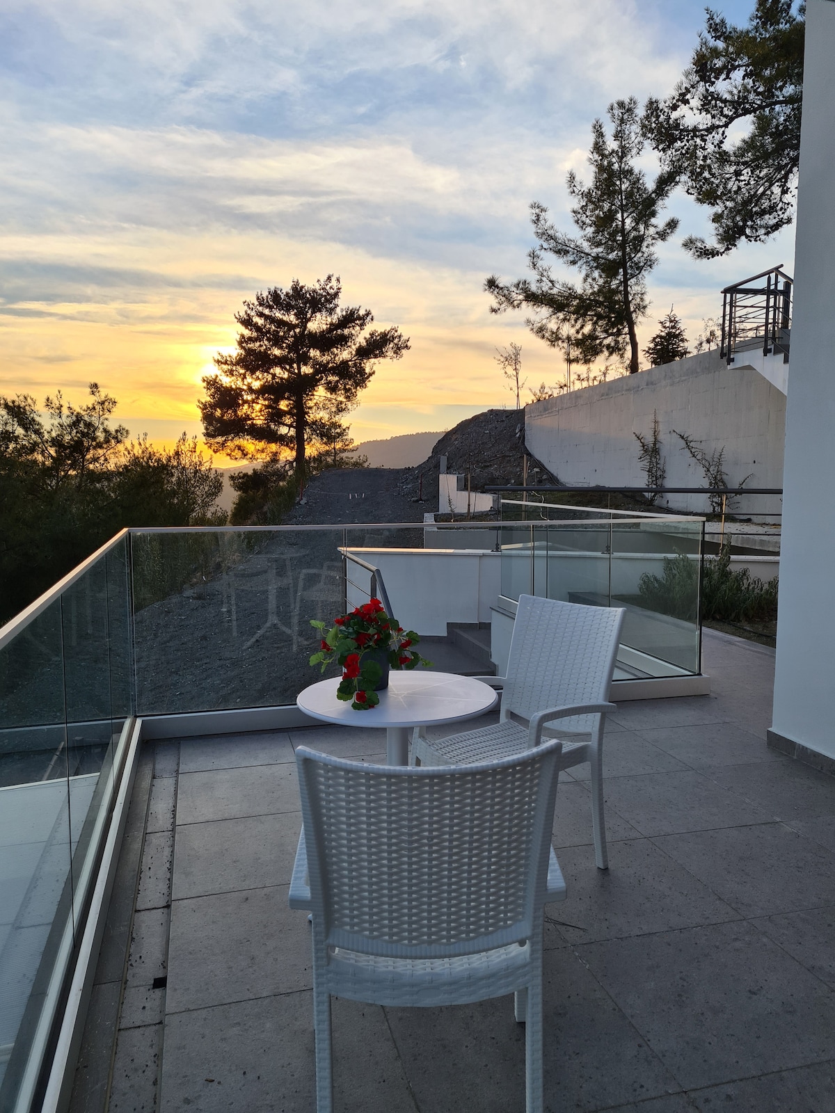 An outdoor balcony area is visible, featuring a small round table accompanied by two chairs. A colorful bouquet sits on the table as the sun sets behind a tree-lined horizon, casting warm hues across the sky.